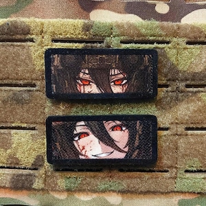 Può includere: Due toppe rettangolari con volti di personaggi in stile anime con occhi rossi e capelli scuri. Le toppe sono nere con retro in velcro. Lo sfondo è un motivo mimetico.