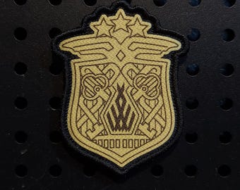 Puede incluir: Un parche bordado en forma de escudo con un diseño en color beige y negro. El emblema muestra tres estrellas en la parte superior, alas estilizadas y dos llaves cruzadas que flanquean un motivo de llama geométrica central, con borde negro.