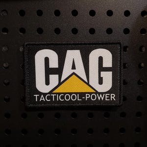 May include: A rectangular embroidered patch with a black border. The centre features the white text CAG with a yellow triangle replacing the letter A. Below this, the white text TACTICOOL-POWER is printed on a black background.