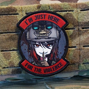 以下が含まれることがあります： 赤色の「I'M JUST HERE FOR THE VIOLENCE」の文字が黒とグレーの背景に刺繍されたパッチ。パッチには、軍用ヘルメットをかぶり、唇に指を当てたアニメスタイルのキャラクターが描かれています。