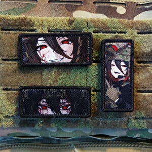 Può includere: Tre toppe rettangolari con disegni di personaggi in stile anime. Le toppe presentano immagini di un personaggio con occhi rossi e capelli scuri. Una toppa mostra un volto sorridente, un'altra mostra gli occhi e la terza mostra un personaggio in uniforme militare.