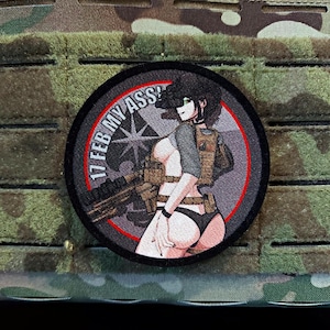 May include: A round patch featuring an anime-style illustration of a woman in tactical gear, holding a weapon. The patch has a red border and the text "17 FEB MY ASS!" in white. The background is a camouflage pattern.