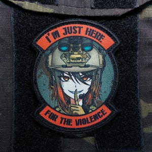 Może przedstawiać: Czarna naszywka z okrągłym wzorem przedstawiającym postać w stylu anime w wojskowym hełmie. Na naszywce znajduje się napis "I'M JUST HERE" nad postacią i "FOR THE VIOLENCE" pod spodem.
