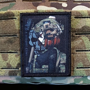 Può includere: Una toppa rettangolare con un'illustrazione in stile anime di una persona in tenuta militare, tra cui un elmetto e un giubbotto tattico. Il personaggio ha i capelli rossi ed è ambientato su uno sfondo scuro. Il testo "SPEED SURPRISE V OF A" è stampato verticalmente.