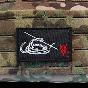 以下が含まれることがあります： 白い蛇が剣に巻き付いた黒い長方形のパッチと赤い悪魔の顔。パッチは白と赤の糸で刺繍されています。背景は迷彩柄です。