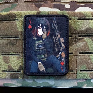 May include: A rectangular patch featuring an anime-style illustration of a person with black hair and red highlights, wearing tactical gear and holding a rifle. The patch includes the Ace of Spades playing card symbol.