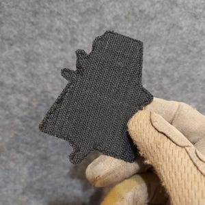 May include: Black hook and loop patch with a textured surface. The patch is shaped like a stylized figure.
