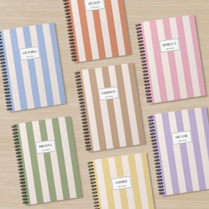 May include: Spiral-bound notebooks with vertical striped covers in various colors, including blue, pink, green, and tan. Each notebook features a white label with a name printed on it. The notebooks are arranged on a light wood surface.