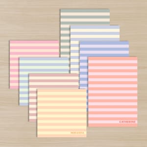 May include: A collection of square notepads with horizontal stripes in pastel colors, including pink, blue, yellow, and green. Two notepads have names printed in the bottom right corner. The notepads are arranged on a light wood surface.