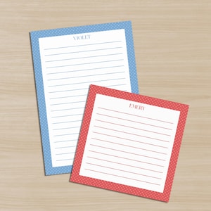 May include: Two lined notepads with polka dot borders. One notepad has a light blue border and the name "VIOLET" at the top. The other notepad has a red border and the name "EMERY" at the top. Both notepads are on a light wood surface.