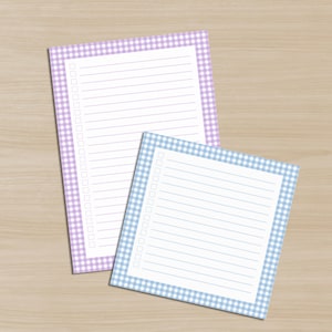 May include: Two notepads with lined pages and a gingham border. One notepad has a light purple border, and the other has a light blue border. The notepads are on a light brown wooden surface. Each notepad has a checklist on the left side.