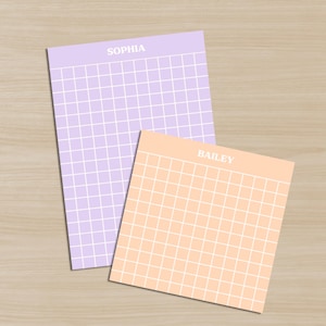 May include: Two square notepads, one lavender and one peach, with a grid pattern and the names "SOPHIA" and "BAILEY" printed at the top. The notepads are on a light wood-grain surface.