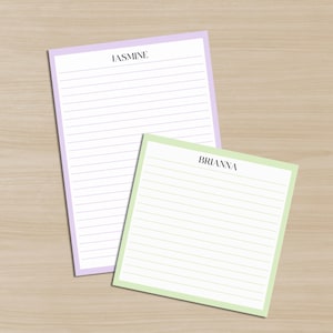 May include: Two lined notepads, one lavender and one light green, rest on a light wood surface. The lavender notepad has the name "JASMINE" printed at the top. The light green notepad has the name "BRIANNA" printed at the top.