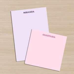 May include: Two personalized notepads, one lavender and one pink, are displayed on a wooden surface. The lavender notepad has the name "MIRANDA" printed in dark purple. The pink notepad has the name "ANASTASIA" printed in a gradient pink.