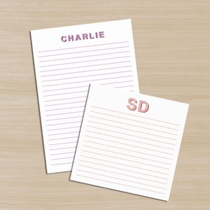 May include: Two lined notepads with the names "CHARLIE" and "SD" printed at the top. The text is in a 3D style. The notepad on top has purple text, and the notepad on the bottom has pink text. The notepads are on a light brown wooden surface.