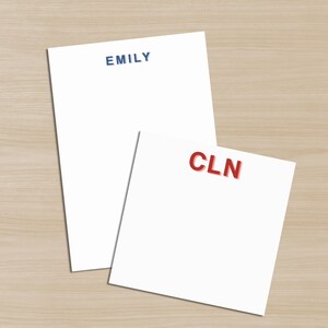 May include: Two white notepads with personalized text. One notepad has the name "EMILY" in navy blue, and the other has the initials "CLN" in red. The notepads are on a light brown wooden surface.