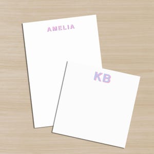 May include: Two white notepads with personalized text. The larger notepad has the name "AMELIA" printed in a gradient of pink and purple. The smaller notepad has the initials "KB" in the same gradient.