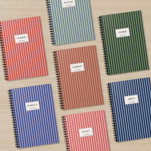 May include: A selection of spiral-bound notebooks with vertical striped covers in various colours, including red, blue, green, and pink. Each notebook has a white label with a name printed on it. The notebooks are arranged on a light wood surface.