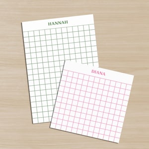 Può includere: Due blocchi per appunti bianchi con motivi a griglia. Uno ha una griglia verde e il nome "HANNAH" in alto. L'altro ha una griglia rosa e il nome "DIANA" in alto. I blocchi per appunti sono su una superficie di legno marrone chiaro.