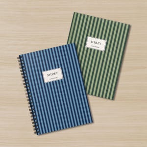 May include: Two spiral-bound notebooks with vertical stripes. One notebook is blue with a label that reads "SYDNEY notes & ideas." The other notebook is green with a label that reads "HAILEY notes & ideas." Both notebooks are on a light wood surface.