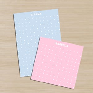 Puede incluir: Dos blocs de notas personalizados, uno azul claro y otro rosa, con lunares blancos. El bloc de notas azul claro tiene el nombre "ELIANA" impreso en la parte superior, mientras que el bloc rosa tiene "ISABELLA".