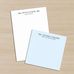 以下が含まれることがあります： 木製の表面に、白と水色の2つのパーソナライズされたメモ帳があります。白いメモ帳には、黒い文字で「DR. OLIVIA LEWIS, MD FAMILY MEDICINE」と書かれています。青いメモ帳には「MS. AMANDA COLLINS SPECIAL EDUCATION TEACHER」と書かれています。