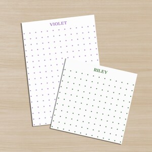Puede incluir: Dos blocs de notas blancos con patrones de puntos. Un bloc de notas tiene puntos morados y la palabra "VIOLET" en la parte superior. El otro bloc de notas tiene puntos verdes y la palabra "RILEY" en la parte superior. Los blocs de notas están sobre una superficie de madera marrón claro.