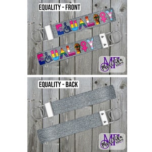 May include: Two keychains with the word "EQUALITY" in rainbow colors and a grey background. The back of the keychain is grey. The keychains are on a wooden surface.