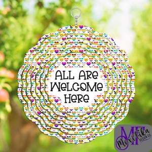 May include: A round, layered decoration with a scalloped edge, featuring a repeating heart pattern in rainbow colors. The center reads "ALL ARE WELCOME HERE" in black text. A small silver ring is at the top for hanging. The background is a blurred green.