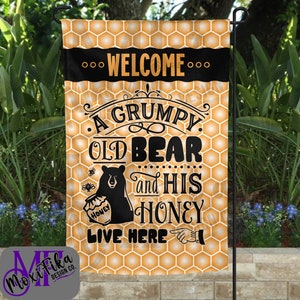 May include: A yellow and black garden flag with a honeycomb pattern. The flag features a black bear holding a jar of honey and the text "...WELCOME... A GRUMPY. OLD BEAR and HIS HONEY LIVE HERE".