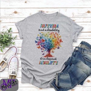 Puede incluir: Una camiseta gris con un árbol arcoíris y mariposas. El texto en la camiseta dice "AUTISM is not a disability it's a different ABILITY".