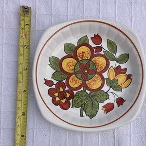 May include: A white ceramic plate with a floral design. The flowers are red, yellow, and green. The plate is 15 cm in diameter.