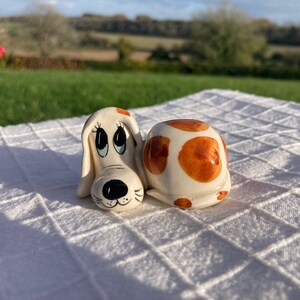 May include: A ceramic dog ornament with a cream base and brown spots. The dog has large eyes, a black nose, and a smiling mouth. The ornament is on a white, textured surface, with a blurred outdoor background.