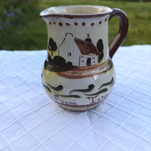 May include: A white ceramic jug with a brown handle and a painted design of a house with a red roof and green trees. The jug has the words "From Torquay" written on the bottom.