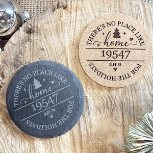 May include: Two round coasters with holiday-themed text. One is dark gray with the words "There's no place like home 19547" and the other is tan with the same text. Both coasters have a Christmas tree graphic.