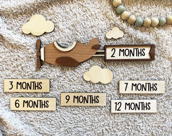 May include: Wooden airplane baby milestone marker with clouds and numbered month cards. The cards include 2 months, 3 months, 6 months, 7 months, 9 months, and 12 months.