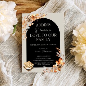 May include: A rectangular invitation card with the text "ADDING S'more LOVE TO OUR FAMILY" in a black arch. The card features a s'mores graphic and is decorated with autumn foliage. The invitation is for a sprinkle in honour of Jenna Ellunan.