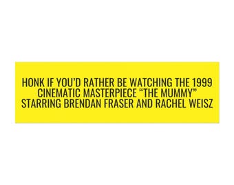 May include: Yellow sign with black text that reads "HONK IF YOU'D RATHER BE WATCHING THE 1999 CINEMATIC MASTERPIECE "THE MUMMY" STARRING BRENDAN FRASER AND RACHEL WEISZ"
