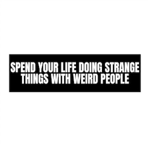 Op de afbeelding: Een zwarte rechthoekige sticker met witte tekst die luidt: "SPEND YOUR LIFE DOING STRANGE THINGS WITH WEIRD PEOPLE."