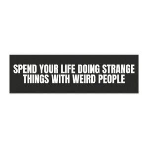 Op de afbeelding: Een zwart rechthoekig bord met witte tekst die luidt: "SPEND YOUR LIFE DOING STRANGE THINGS WITH WEIRD PEOPLE."
