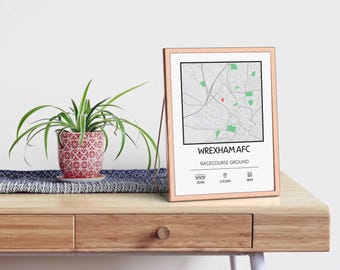 Può includere: Una mappa incorniciata del Racecourse Ground del Wrexham AFC, con uno spillo rosso che segna la posizione dello stadio. La mappa è stampata su carta bianca con testo nero e aree verdi che rappresentano i parchi. La cornice è di colore rame chiaro.