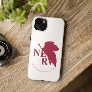 May include: White phone case featuring a burgundy leaf design with the letters "NERV" and the text "GOD'S IN HIS HEAVEN, ALL'S RIGHT WITH THE WORLD." The case is on a wooden surface with a camera and plant in the background.