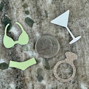 May include: A set of four paper confetti cutouts in various shapes, including a bikini, a martini glass, a ring, and a sparkling diamond. The confetti is light green, white, and silver.