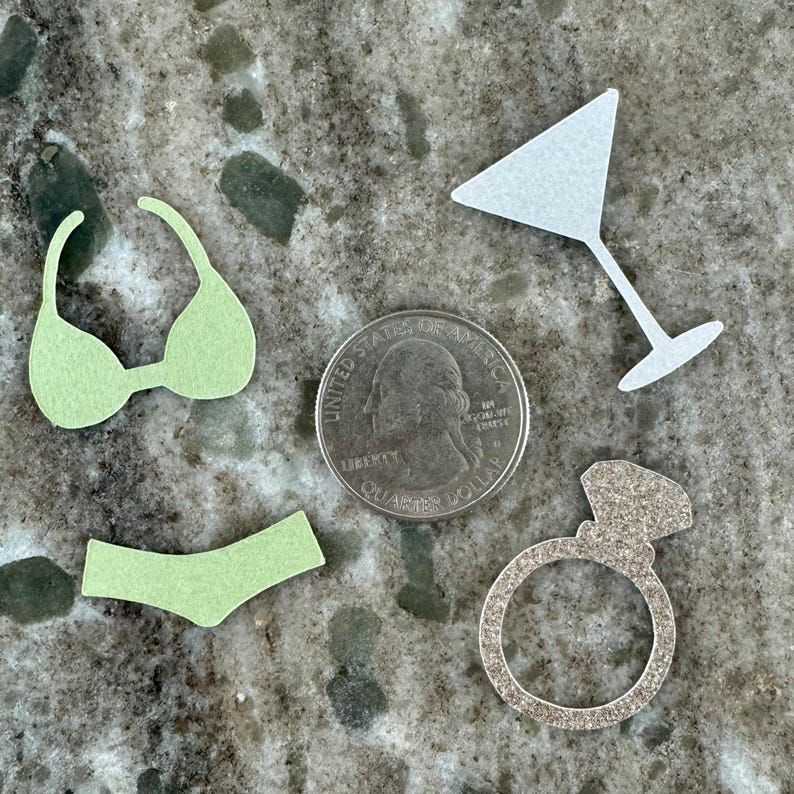 May include: A set of four paper confetti cutouts in various shapes, including a bikini, a martini glass, a ring, and a sparkling diamond. The confetti is light green, white, and silver.