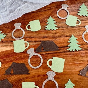 May include: Green and brown paper cutouts of camping themed items, including a tent, a mug, a pine tree, and a ring. The ring is silver glitter.