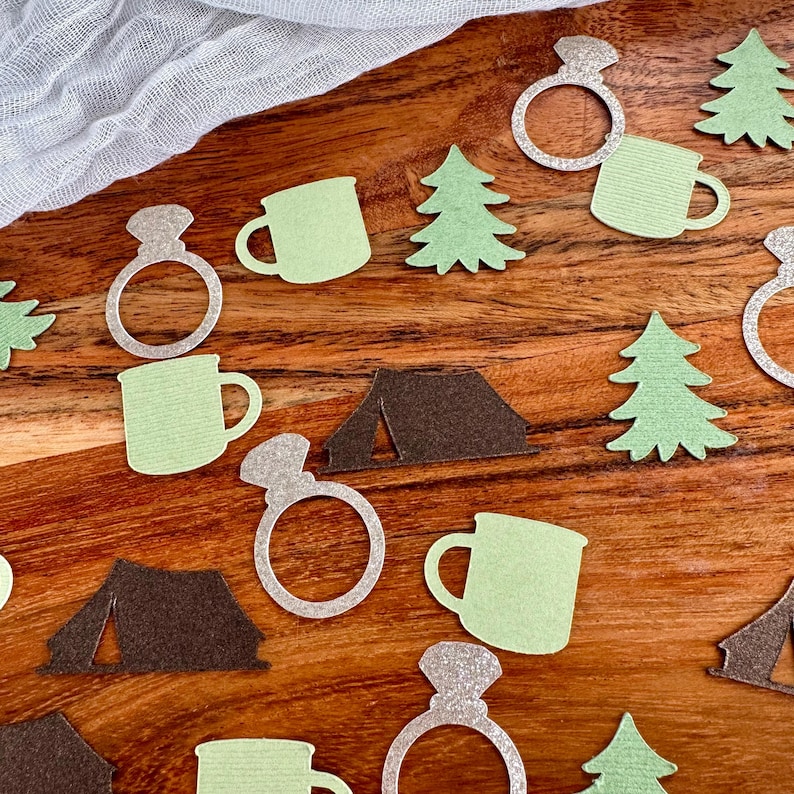 May include: Green and brown paper cutouts of camping themed items, including a tent, a mug, a pine tree, and a ring. The ring is silver glitter.