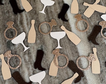 Può includere: Una collezione di coriandoli per feste con forme fustellate che includono bottiglie di champagne beige, coppe da champagne bianche testurizzate, stivali da cowboy neri e anelli di fidanzamento dorati glitterati sparsi su una superficie.