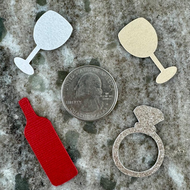 May include: A set of paper cutouts for a wedding or engagement party. The set includes a wine bottle, two wine glasses, and a ring with a diamond. The cutouts are made of different colors of paper, including red, white, and beige.