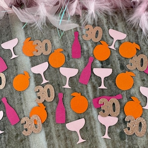 May include: A festive arrangement of party confetti, including pink champagne glasses, orange fruit shapes, and pink bottles. Glittery gold "30" cutouts are scattered throughout. Pink feathers adorn the top, creating a celebratory theme.