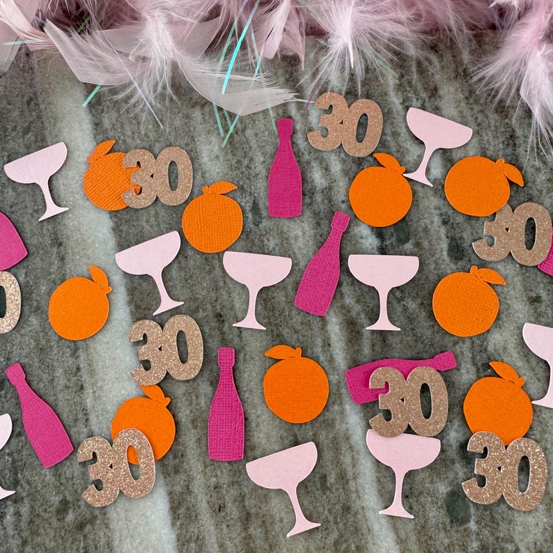 May include: A festive arrangement of party confetti, including pink champagne glasses, orange fruit shapes, and pink bottles. Glittery gold "30" cutouts are scattered throughout. Pink feathers adorn the top, creating a celebratory theme.